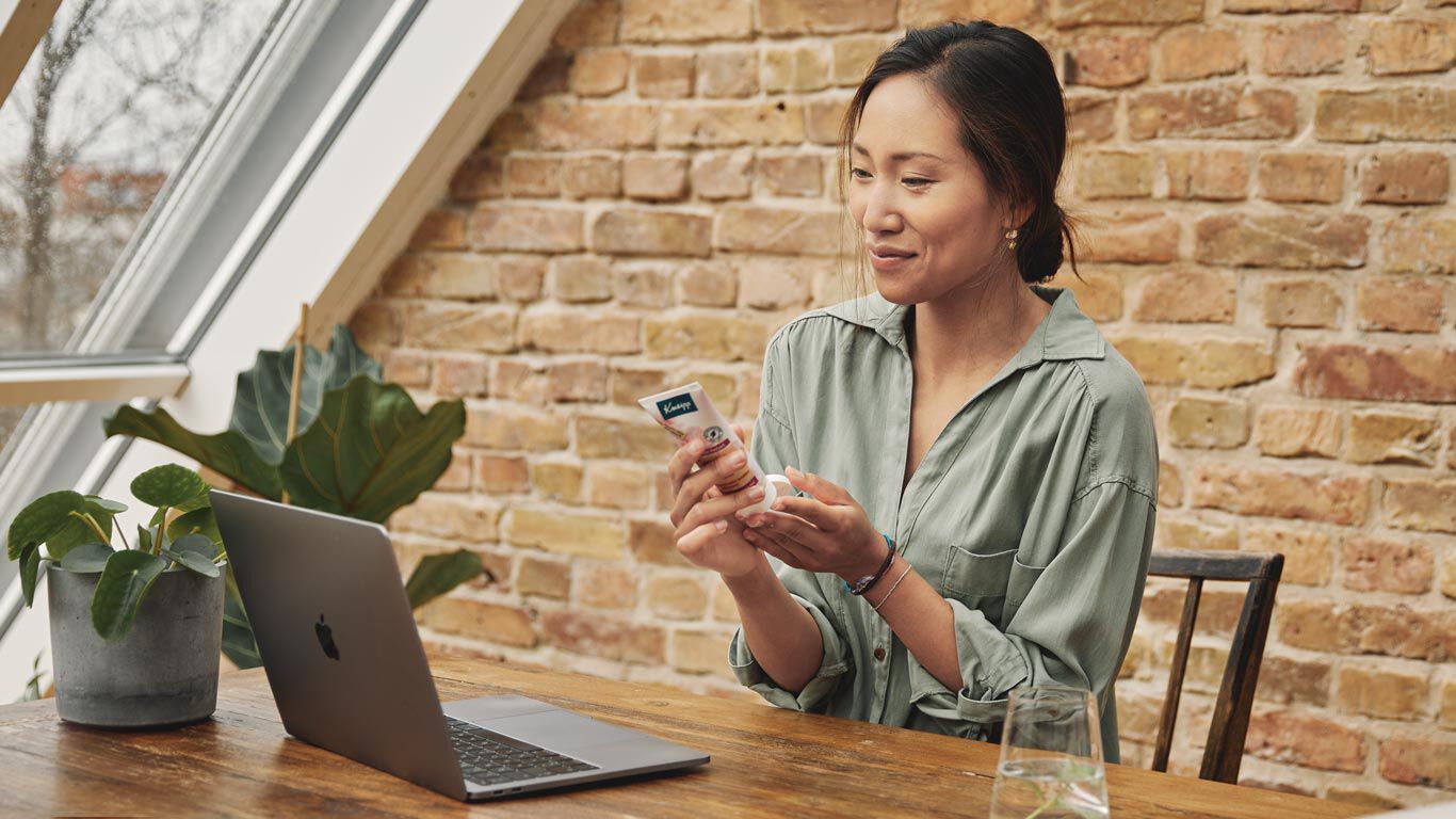 Frau sitzt am Tisch vor ihrem Laptop und cremt sich die Hände ein.